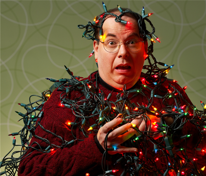 Picture is of a man wrapped up in Christmas lights looking frightened. His mouth is held open, and he is wearing glasses.  